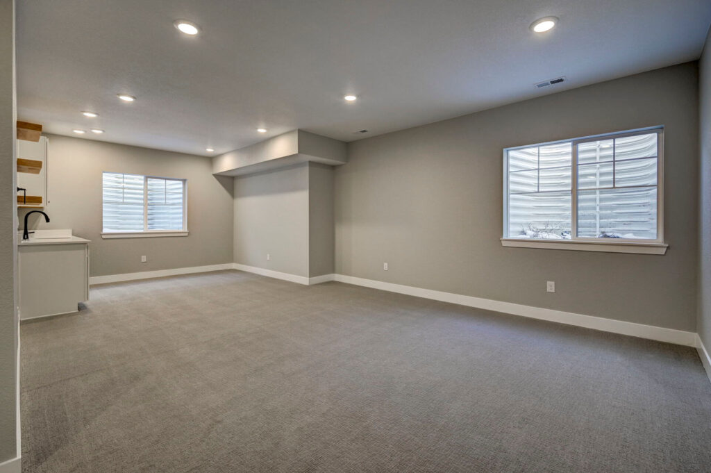 Large open finished basement room with two egress windows and a small sink area to the left.