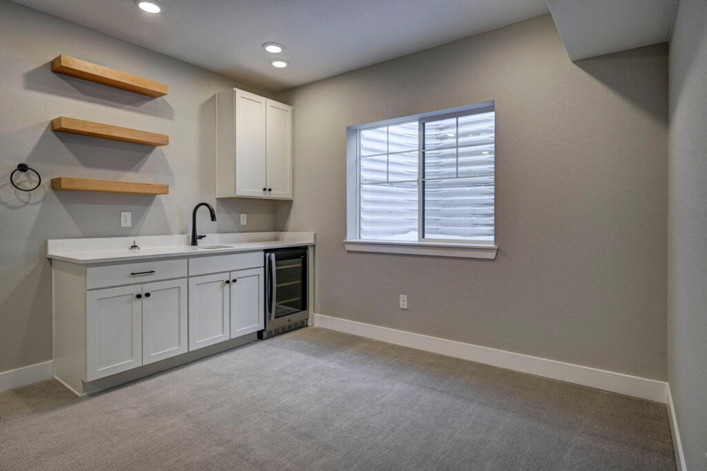 Small kitchenette area with white cabinets, countertop sink, floating wooden shelves, and a wine fridge.