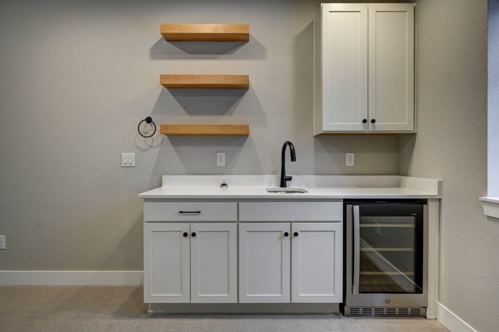 Close-up of a wet bar with white cabinets, small round sink, black faucet, three wooden floating shelves and wine fridge.