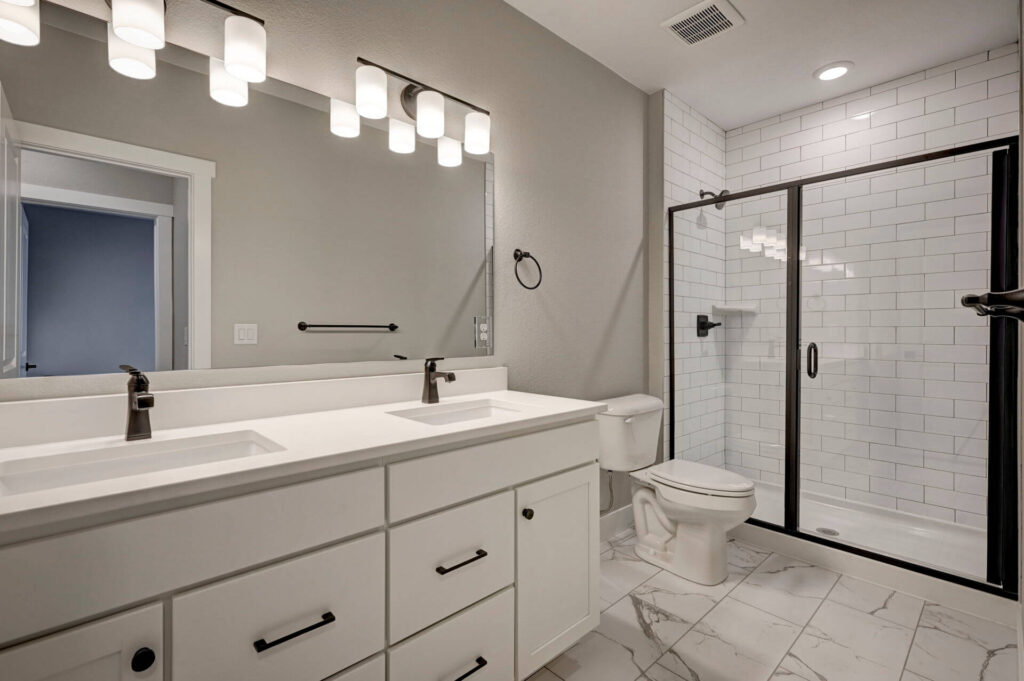 Bright bathroom with double sink vanity, large mirror, toilet, and glass-enclosed tiled shower.