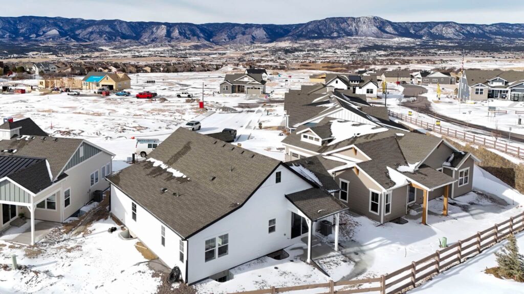 Oblique aerial panorama of a row of houses on snowy lots with mountains and open fields beyond.