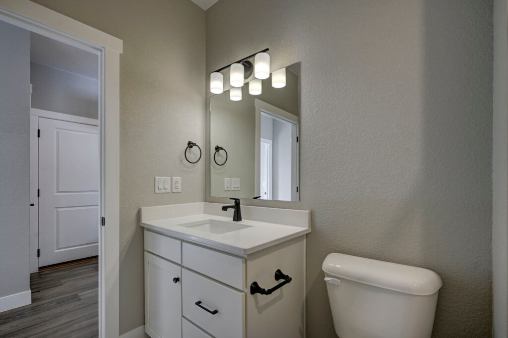 Vanity area with white cabinet, mirror, black faucet and light fixture next to a toilet.