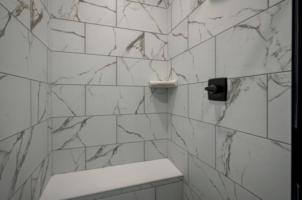 Close-up of shower interior with marble-look tiles, soap shelf and dark faucet control.