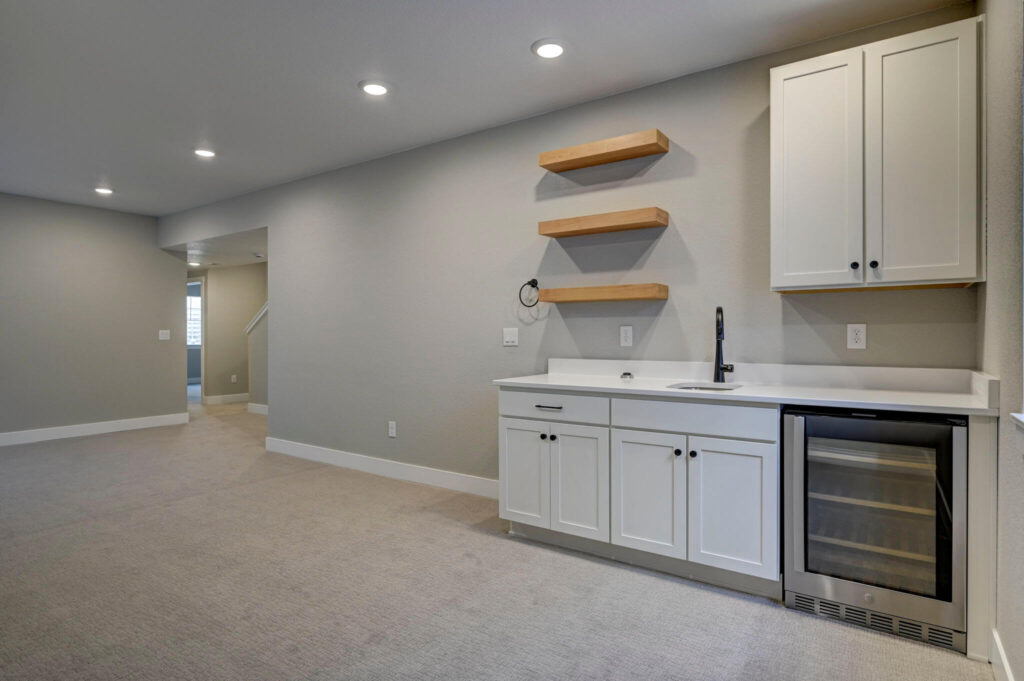 Long view of a finished room with a wet bar, white cabinets, floating shelves, and recessed ceiling lights.