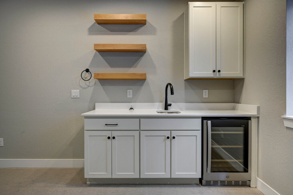 Close-up of a white wet bar with a sink, black faucet, three wooden floating shelves, and a glass-front wine fridge.