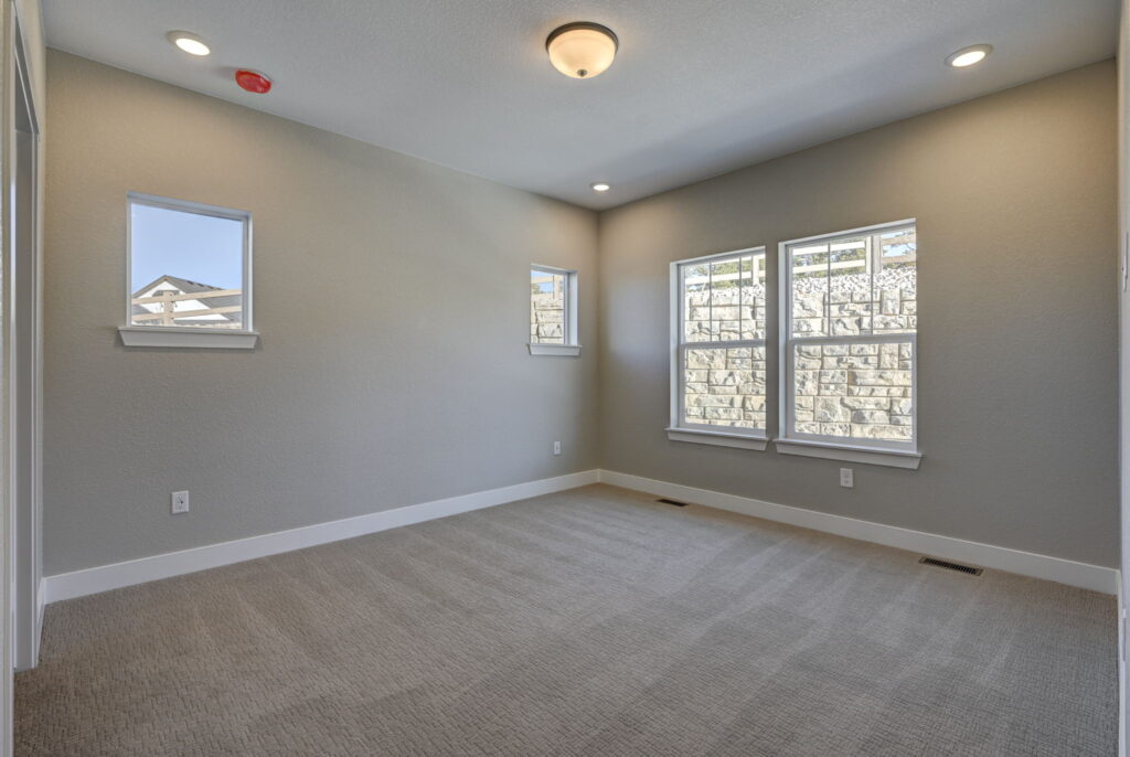 Empty carpeted room with three windows and neutral gray-beige walls under ceiling lights.