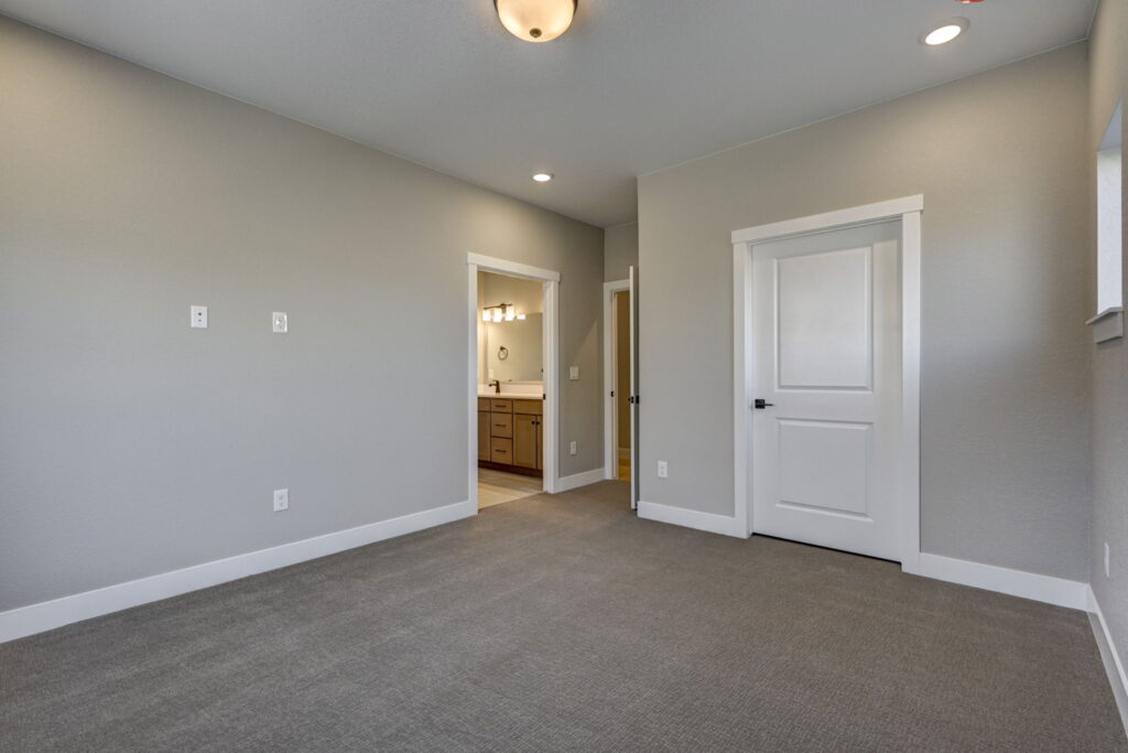 Empty bedroom looking toward bathroom entrance and closed closet door, with wall-mounted lighting in bathroom.