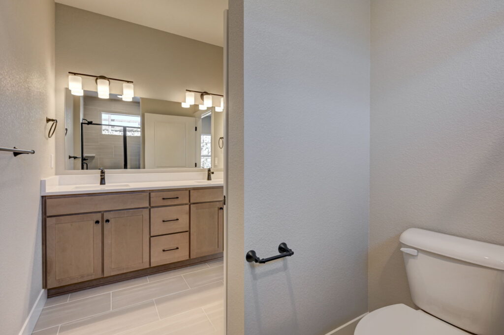 Toilet alcove adjacent to a double-sink vanity seen through a partial wall
