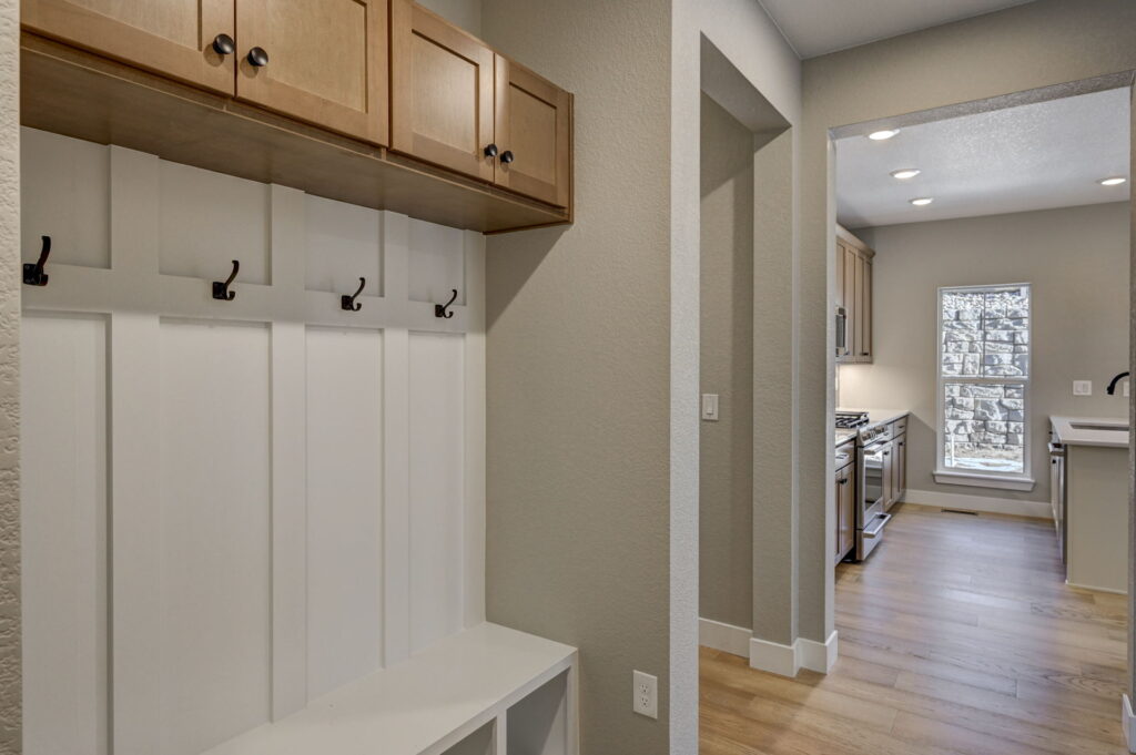 Small entry mudroom with white board-and-batten bench, hooks, upper wood cabinets and view into a kitchen.