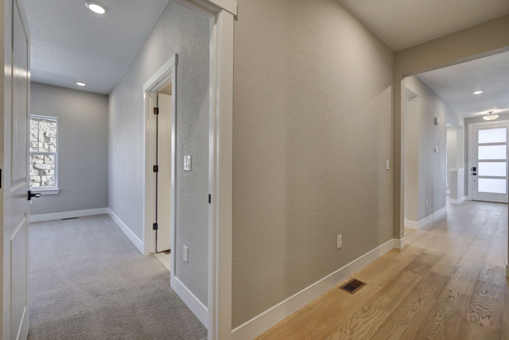 Interior hallway with light wood floors, open doorway to a carpeted room and a front door at the far end.