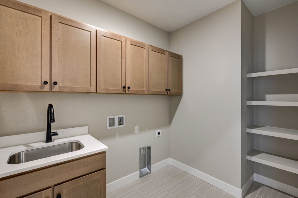 Laundry room sink, upper cabinets and shelving with utility hookups on tiled floor.