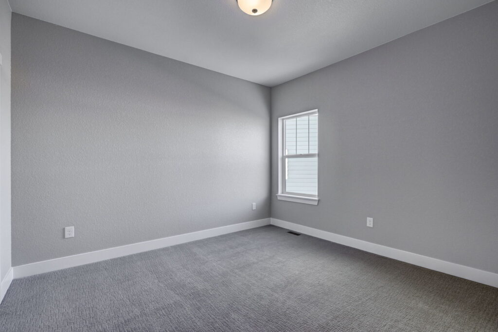 Empty small bedroom with gray carpet, light gray walls, white baseboards, and a single window.