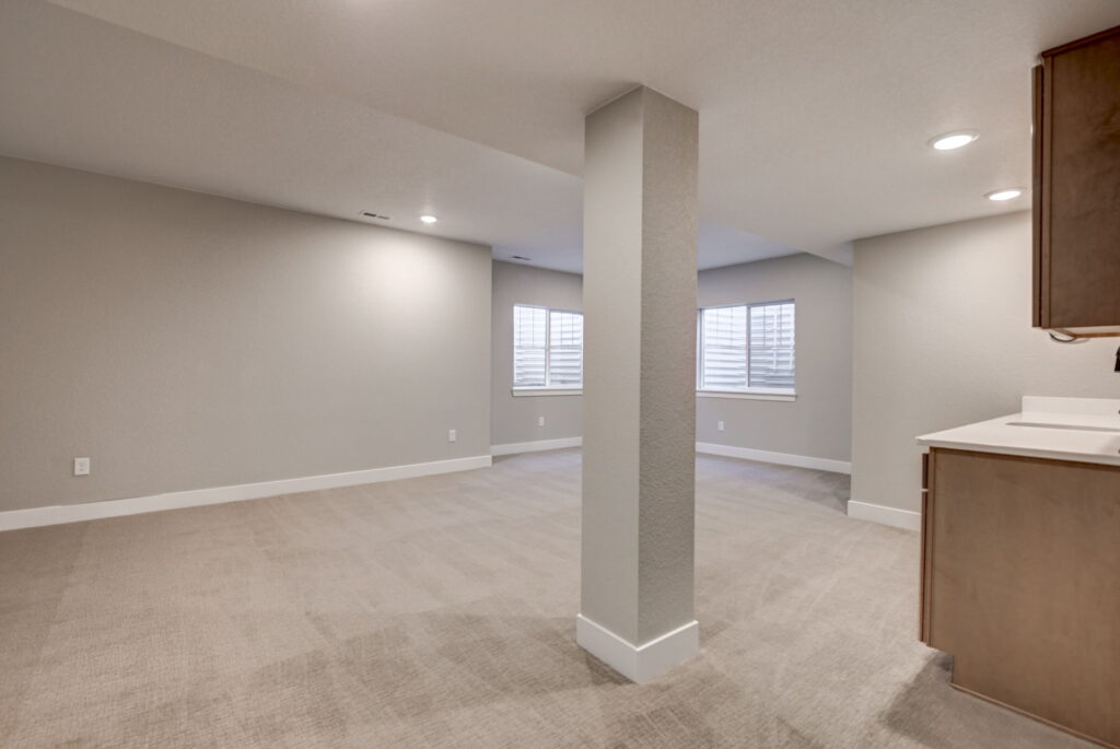 Large open finished basement room with carpet, central support column, and small kitchenette.