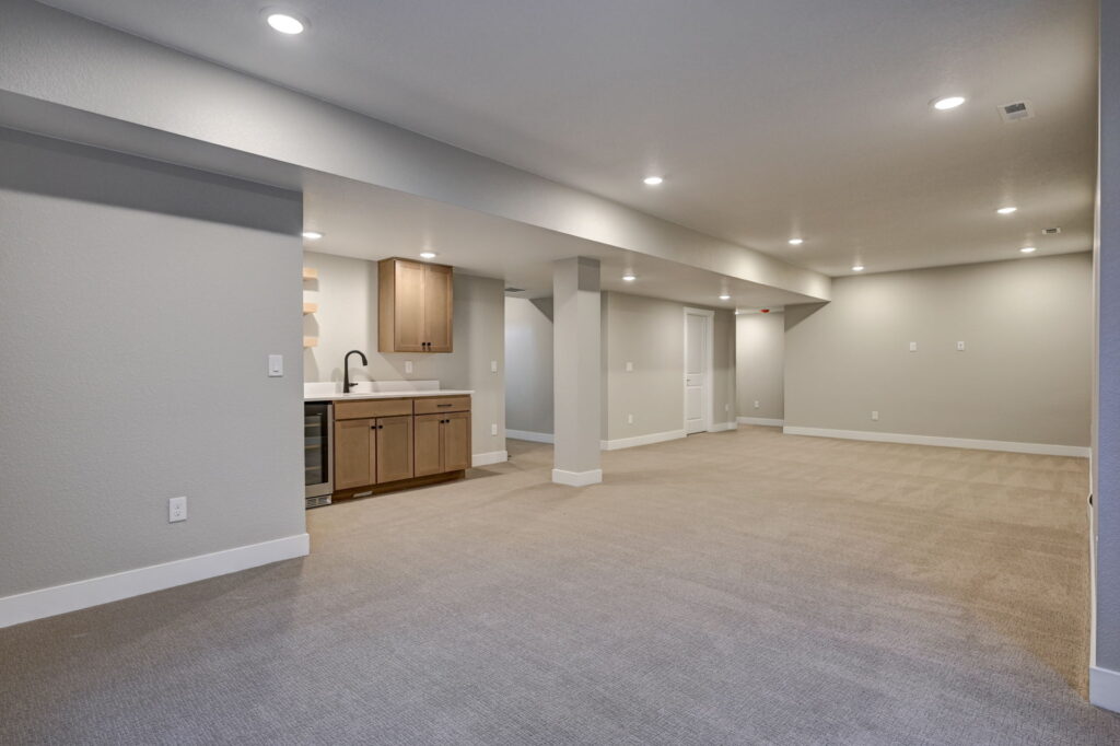 Wide view of a large finished basement with wet bar at one end, support columns and recessed lights.