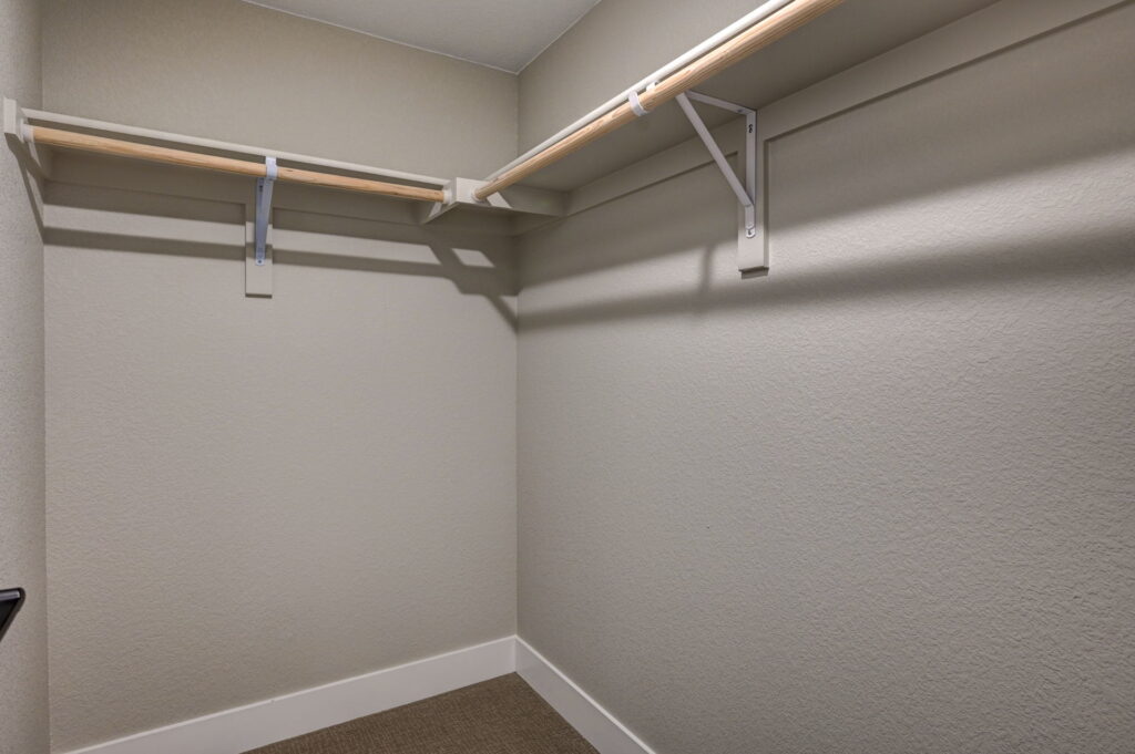Small walk-in closet with two wooden hanging rods and upper shelf on beige walls.