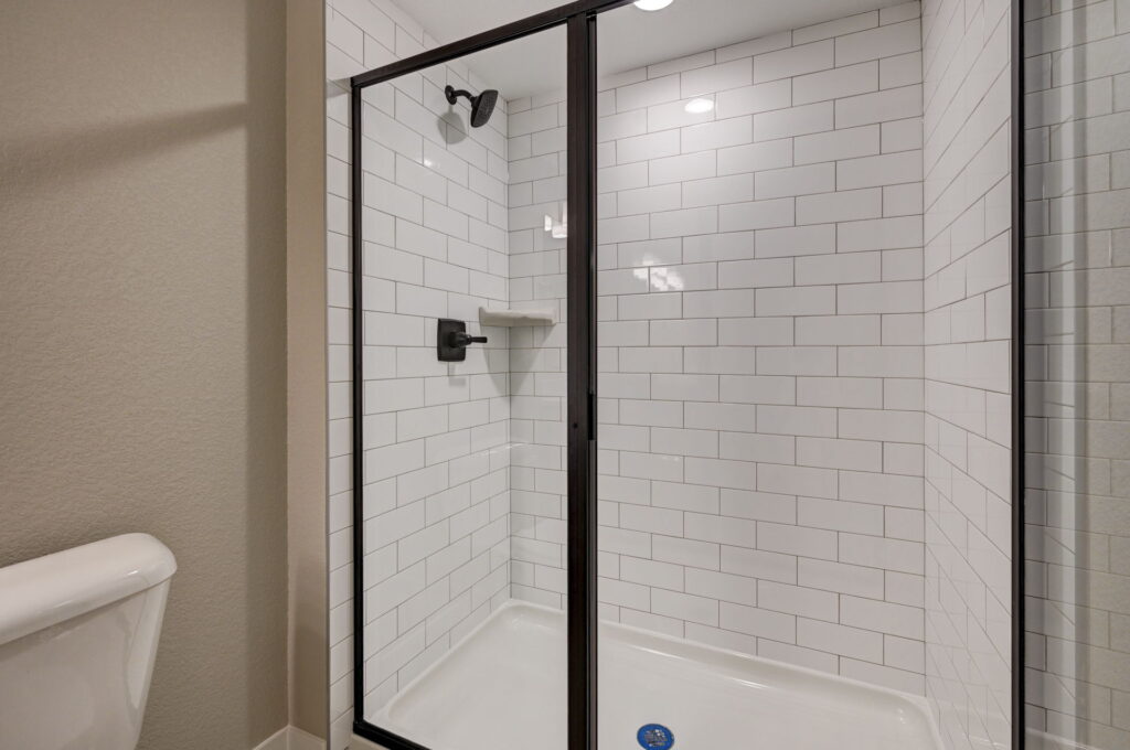 Glass-enclosed shower with black metal frame, white subway tile walls and dark fixtures.