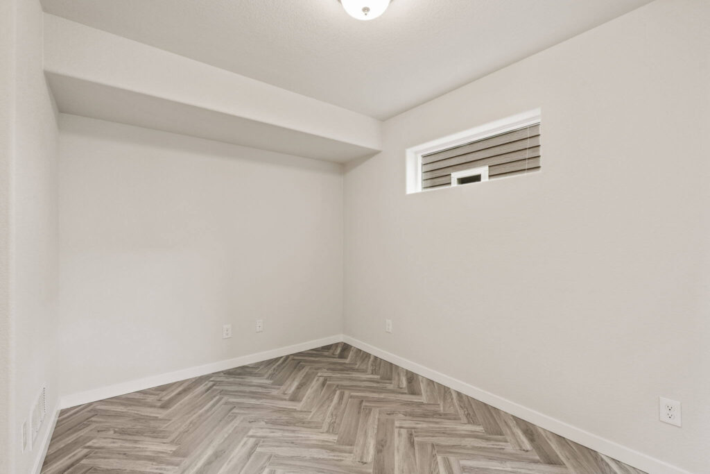 Corner of a small empty room with herringbone-pattern flooring and a narrow high window.