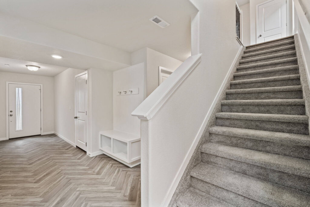 Entry foyer with carpeted staircase and built-in bench with hooks, light herringbone-look flooring.