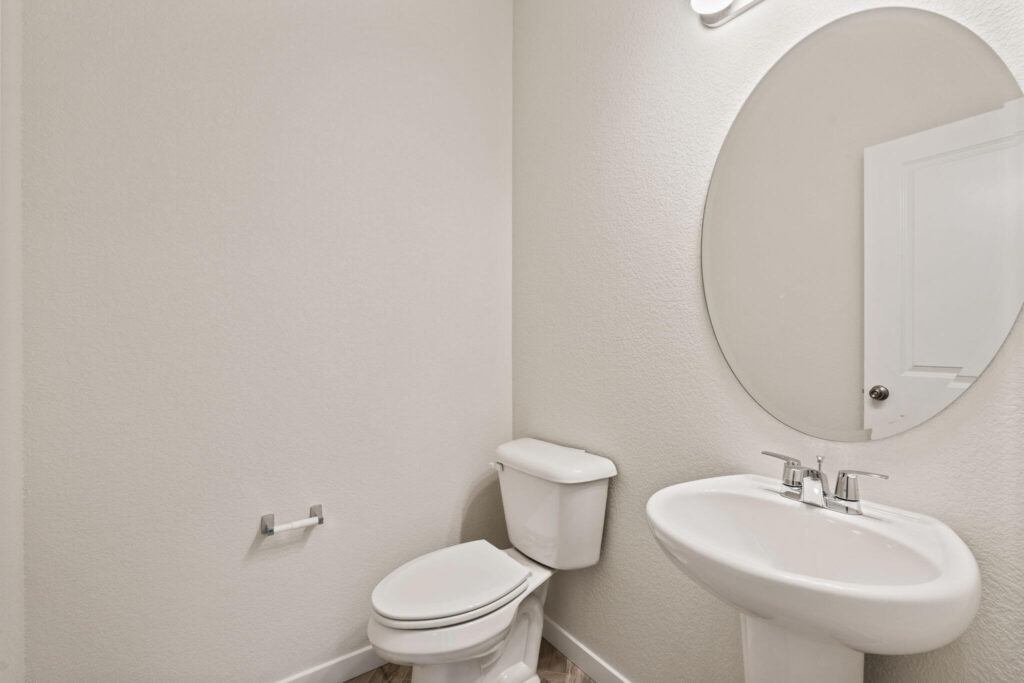 Powder room with pedestal sink, oval mirror, and a toilet against a plain light-colored wall.