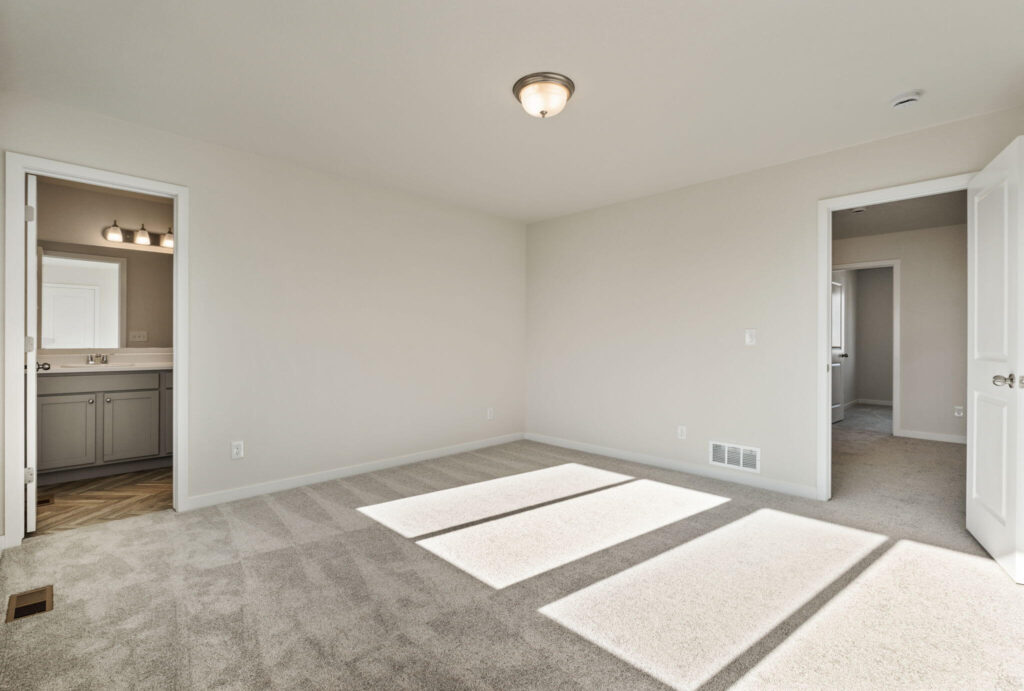 Empty carpeted bedroom with open doorway to a bathroom and sunlight casting rectangular patterns on the floor.