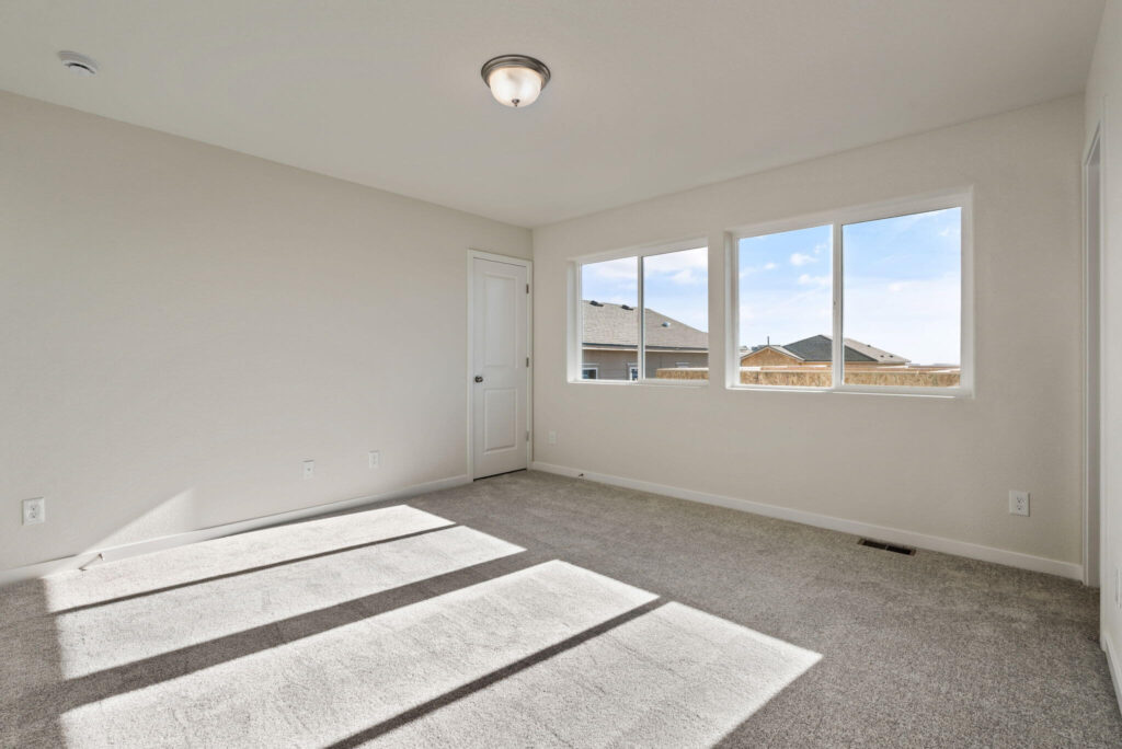 Room with three large windows overlooking neighboring roofs and sunlight falling across a carpeted floor.