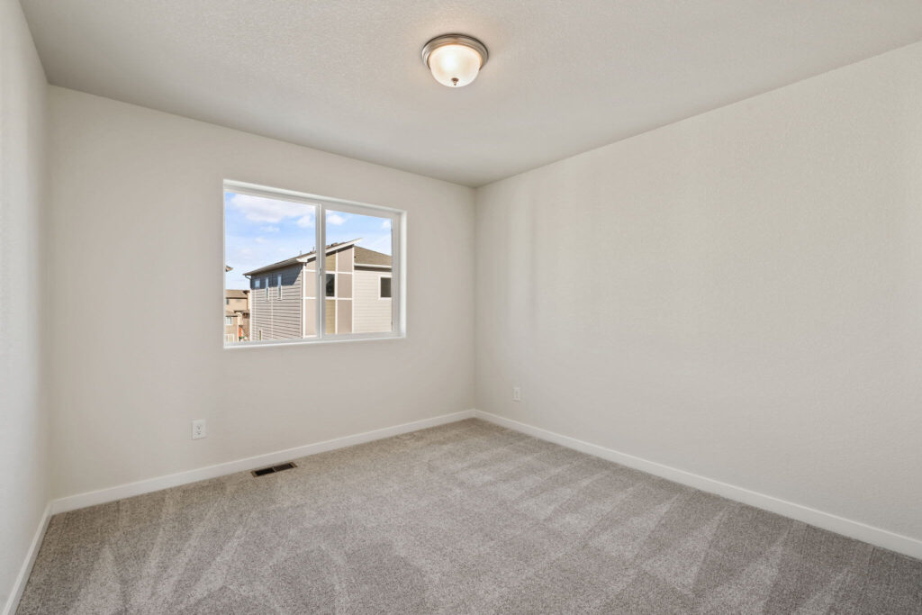 Empty carpeted bedroom with single window showing neighboring house and ceiling light fixture.