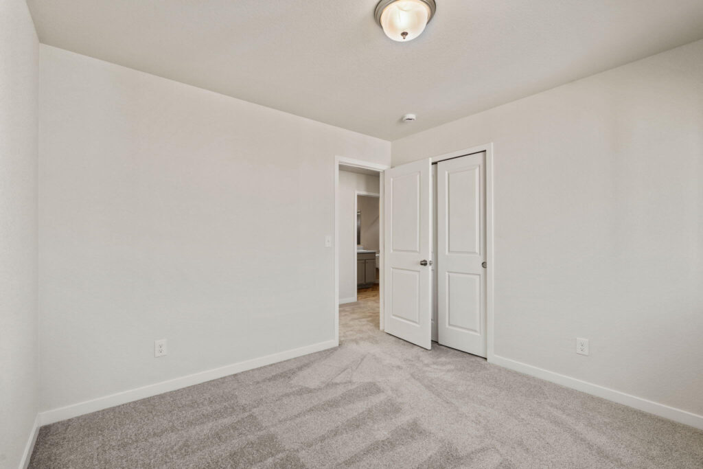 Empty bedroom viewed toward an open doorway and a closet with double doors, neutral carpet and walls.