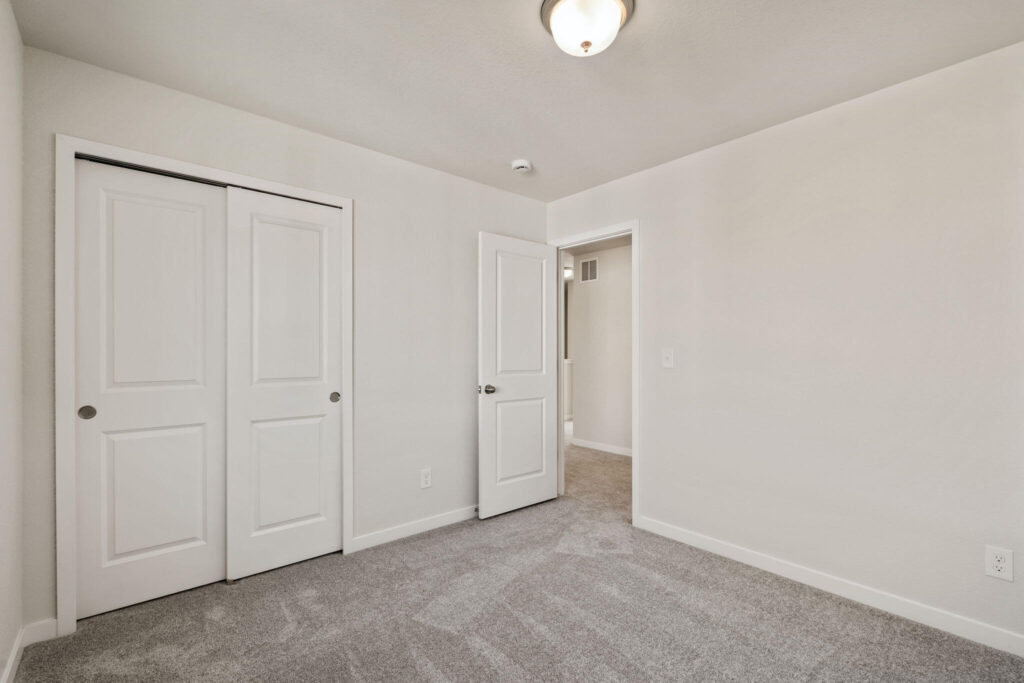 Small empty bedroom with gray carpet, white walls, sliding closet doors and ceiling light.