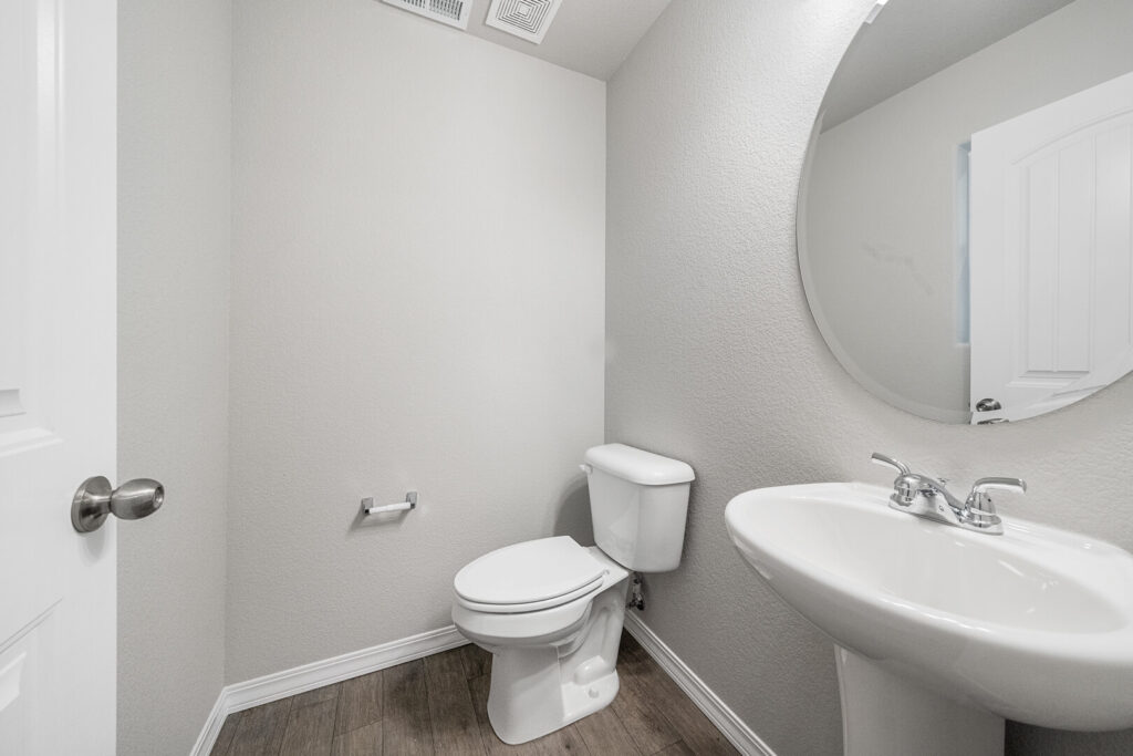 Small half bathroom with pedestal sink, round mirror, and toilet on wood-look flooring.