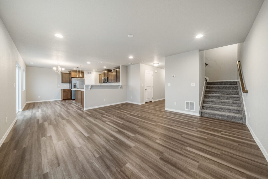 Open-plan living area with wood-look floors, neutral walls, kitchen at rear and carpeted stairs.