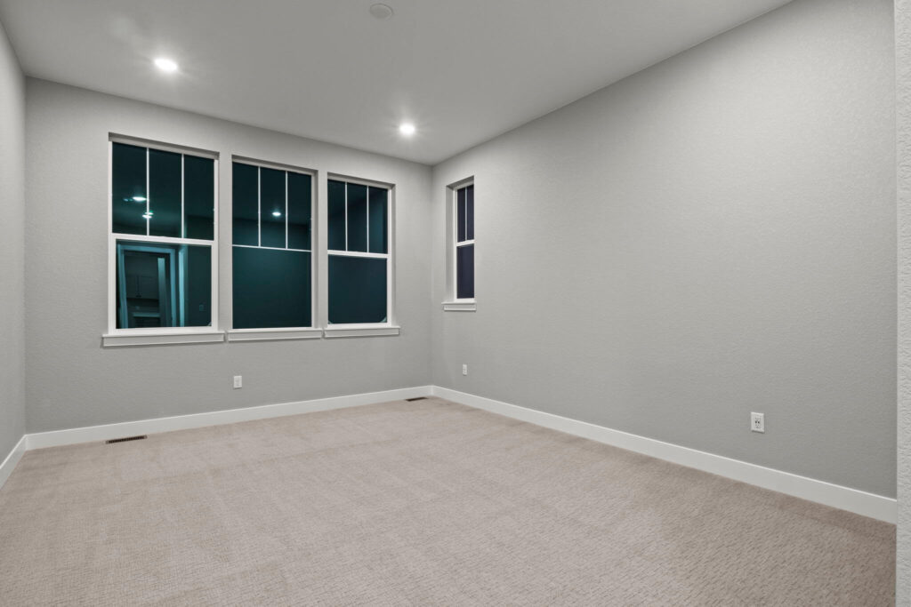 Empty carpeted room with three large windows and recessed ceiling lights.