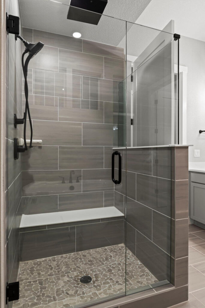 Glass walk-in shower with bench, pebble-pattern floor and large rectangular gray wall tiles.