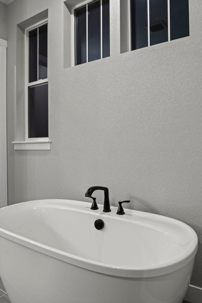Freestanding bathtub beneath two tall windows with black faucet and neutral-colored wall.