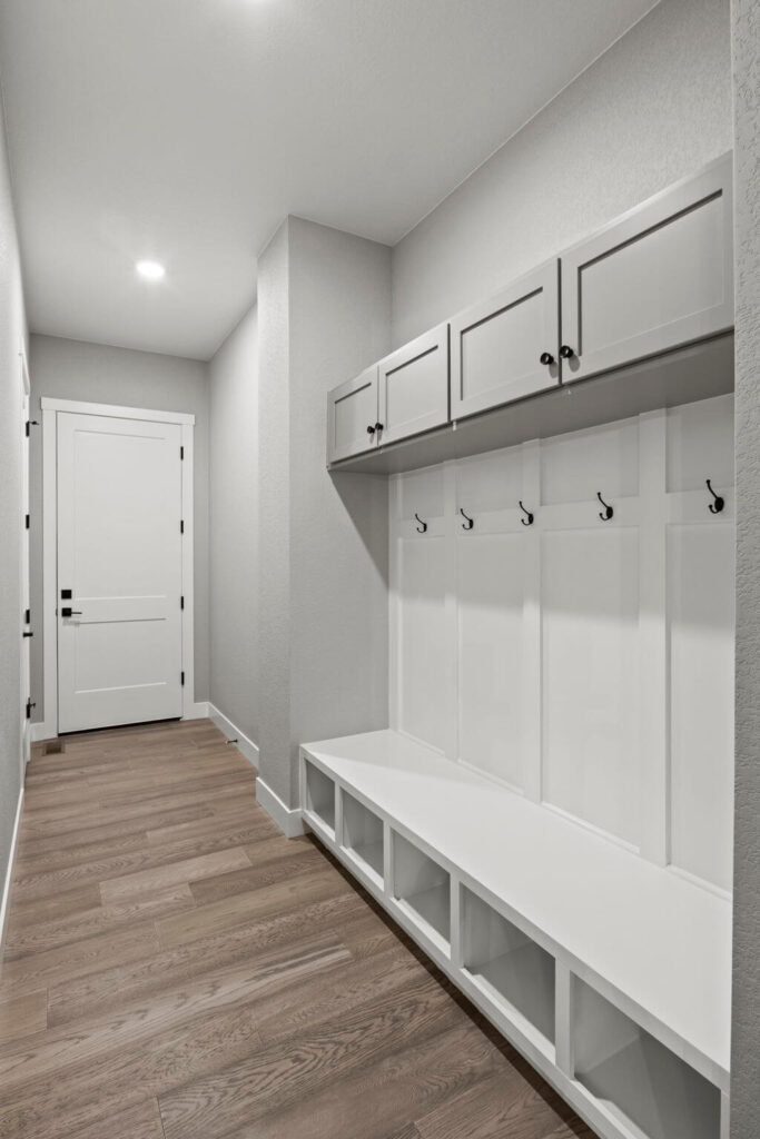Narrow mudroom with built-in bench cubbies, overhead cabinets, and coat hooks along a paneled wall.