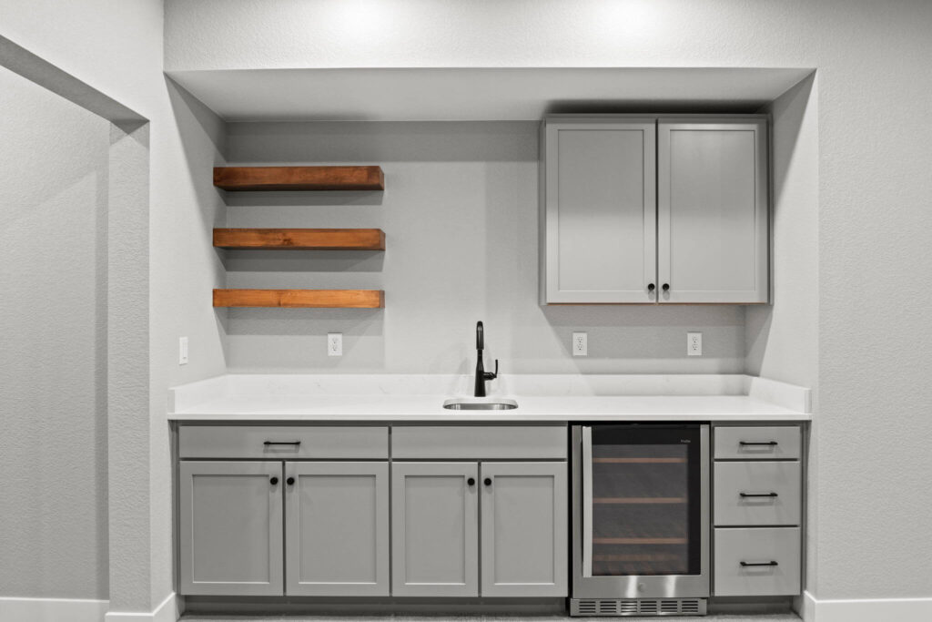 Built-in wet bar with gray cabinets, white countertop, wood floating shelves, and small wine fridge.
