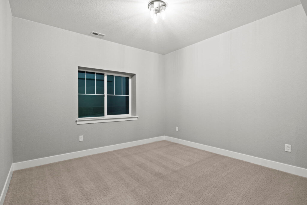 Empty carpeted room with a single window and overhead light, neutral gray walls.