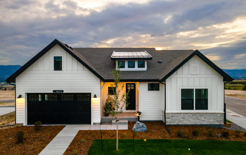 Evening exterior of a white modern farmhouse with lit porch, black garage door, and dormer window.