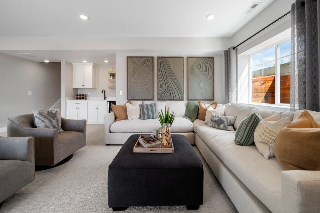 Bright finished basement seating area with L-shaped cream sofa, gray swivel chairs, and black ottoman