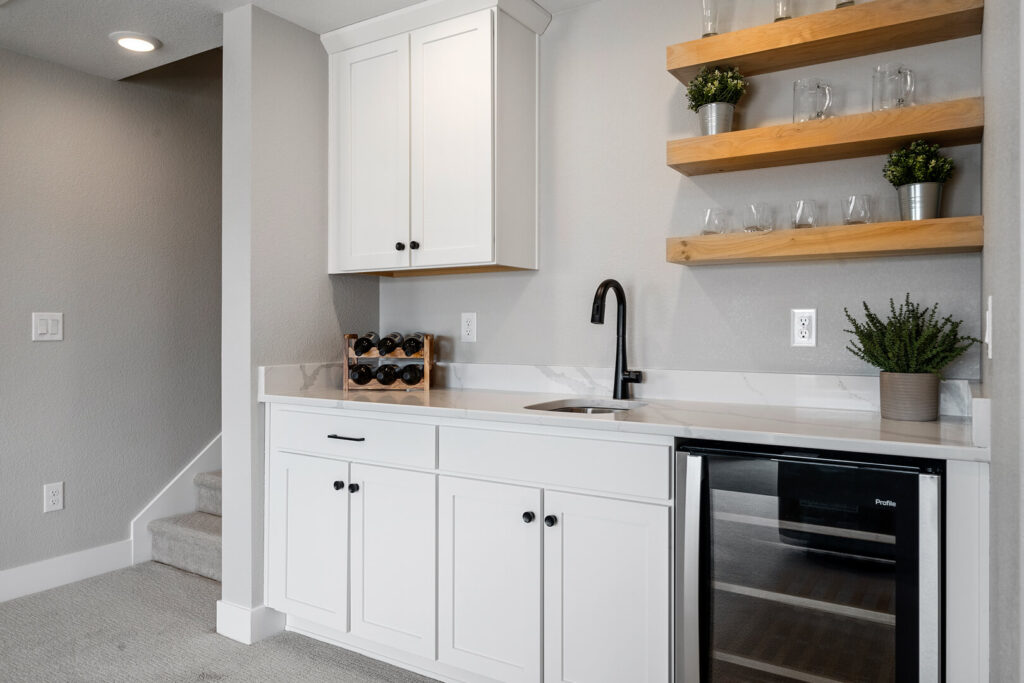 Small kitchenette or wet bar with white cabinets, a sink, open wooden shelves, and a beverage fridge.