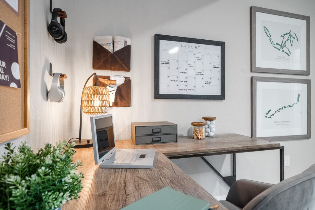 Corner desk workspace with laptop, calendar board, framed art, and lamp on the wall.