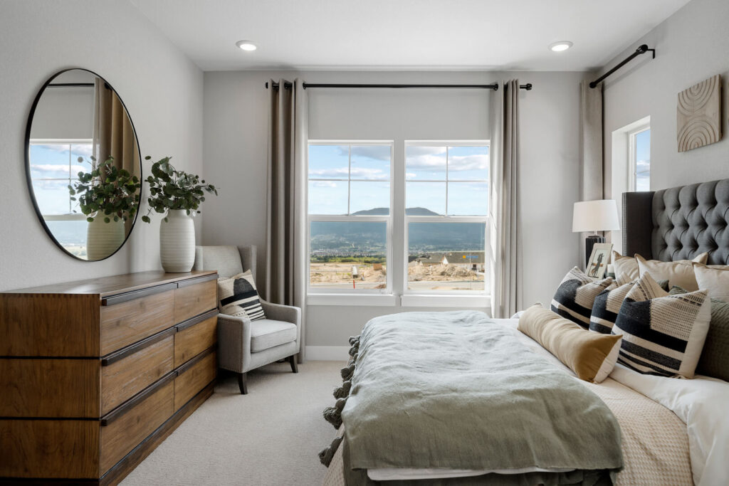 Well-lit bedroom with bed, dresser, accent chair and twin windows showing landscape beyond.