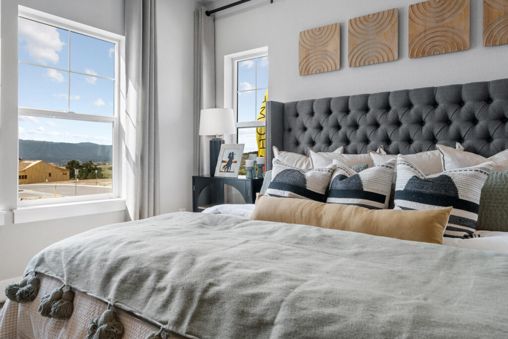 Bed with tufted headboard and layered pillows beside a window overlooking distant hills.