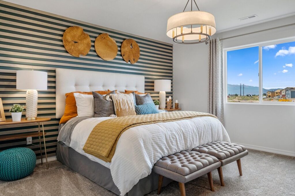 Bed with tufted bench at foot, striped accent wall and window showing distant houses and sky.