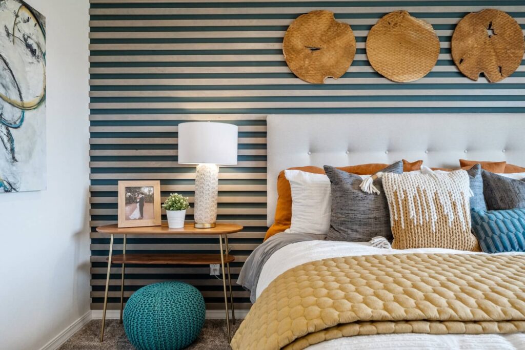 Bed with textured striped accent wall, decorative woven wall art, side table with lamp and knit pouf.