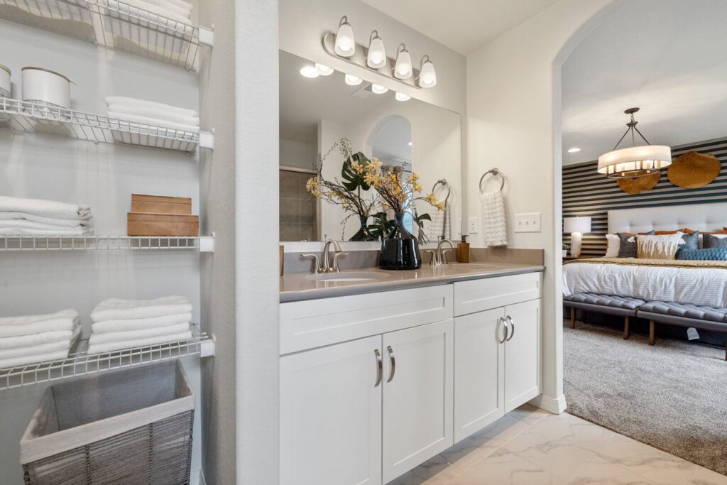 Vanity area with double sinks, mirror, shelving with towels, and view into the bedroom.
