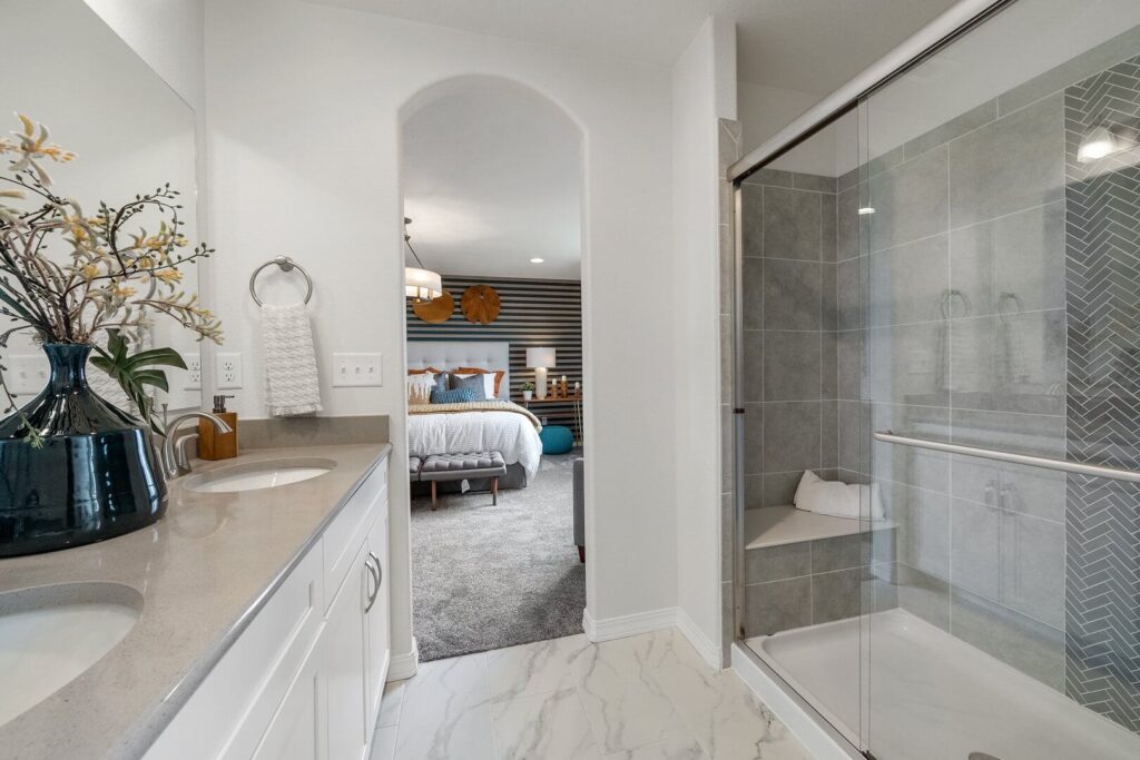 Bathroom with double vanity, marble-look floor and glass-enclosed shower opening toward a bedroom.