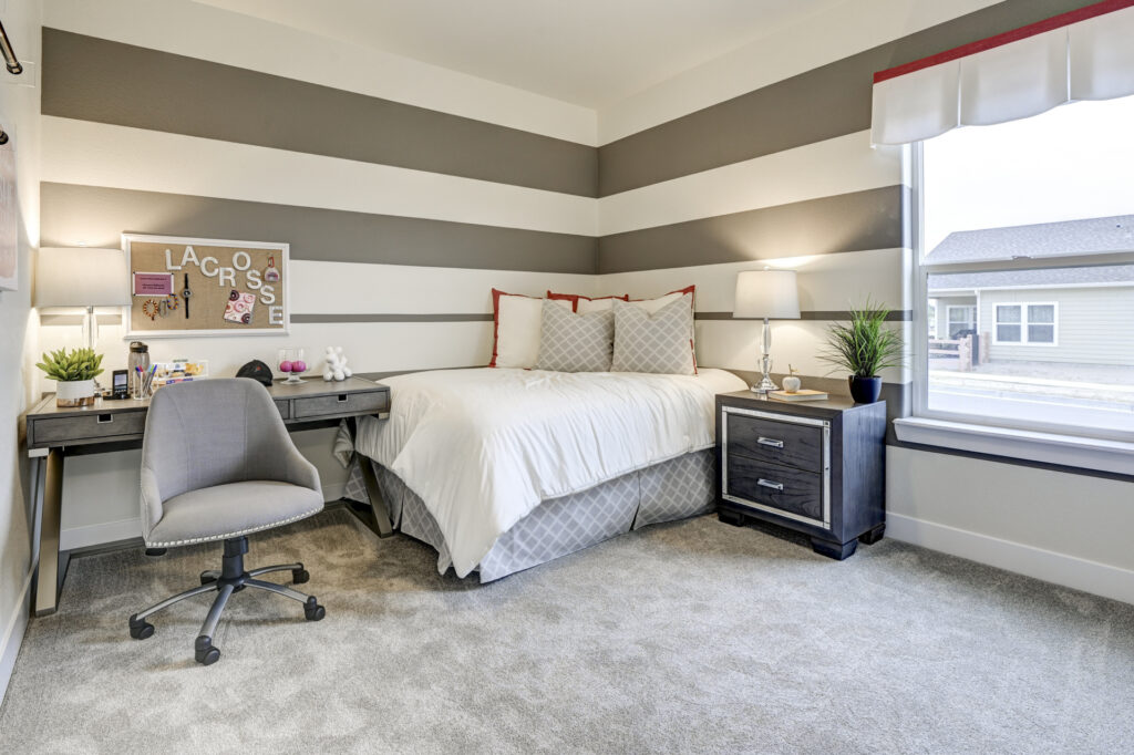 Bedroom with striped accent wall, bed, desk, and rolling office chair near a window.