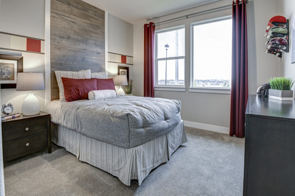 Small bedroom with wood-paneled headboard, gray bedding, red accent pillows, and window with red curtains.