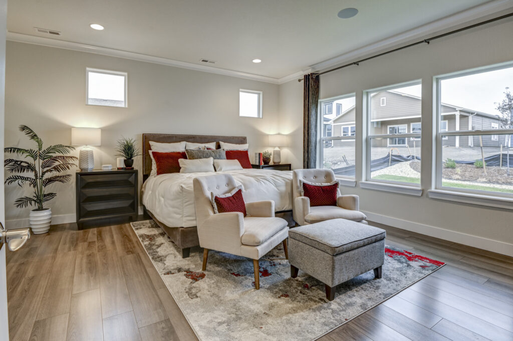 Staged bedroom with bed, two armchairs, ottoman, and large windows.