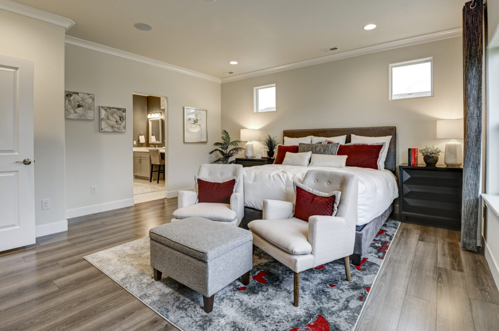 Staged bedroom with two armchairs, ottoman, rug and bed with red accent pillows near doorway.